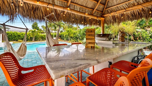 An outdoor dining area with a thatched roof, a pool, and lush greenery in the background. - Eccentric Home With Pool Tiki hut Outdoor Kitchen (Pompano Beach)