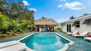 A swimming pool with lounge chairs, a thatched-roof structure, and lush greenery. - Eccentric Home With Pool Tiki hut Outdoor Kitchen (Pompano Beach)