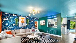 A modern living room with a sofa, coffee table, and patterned rug. A kitchen area is visible with bar stools and a neon sign. - Eccentric Home With Pool Tiki hut Outdoor Kitchen (Pompano Beach)