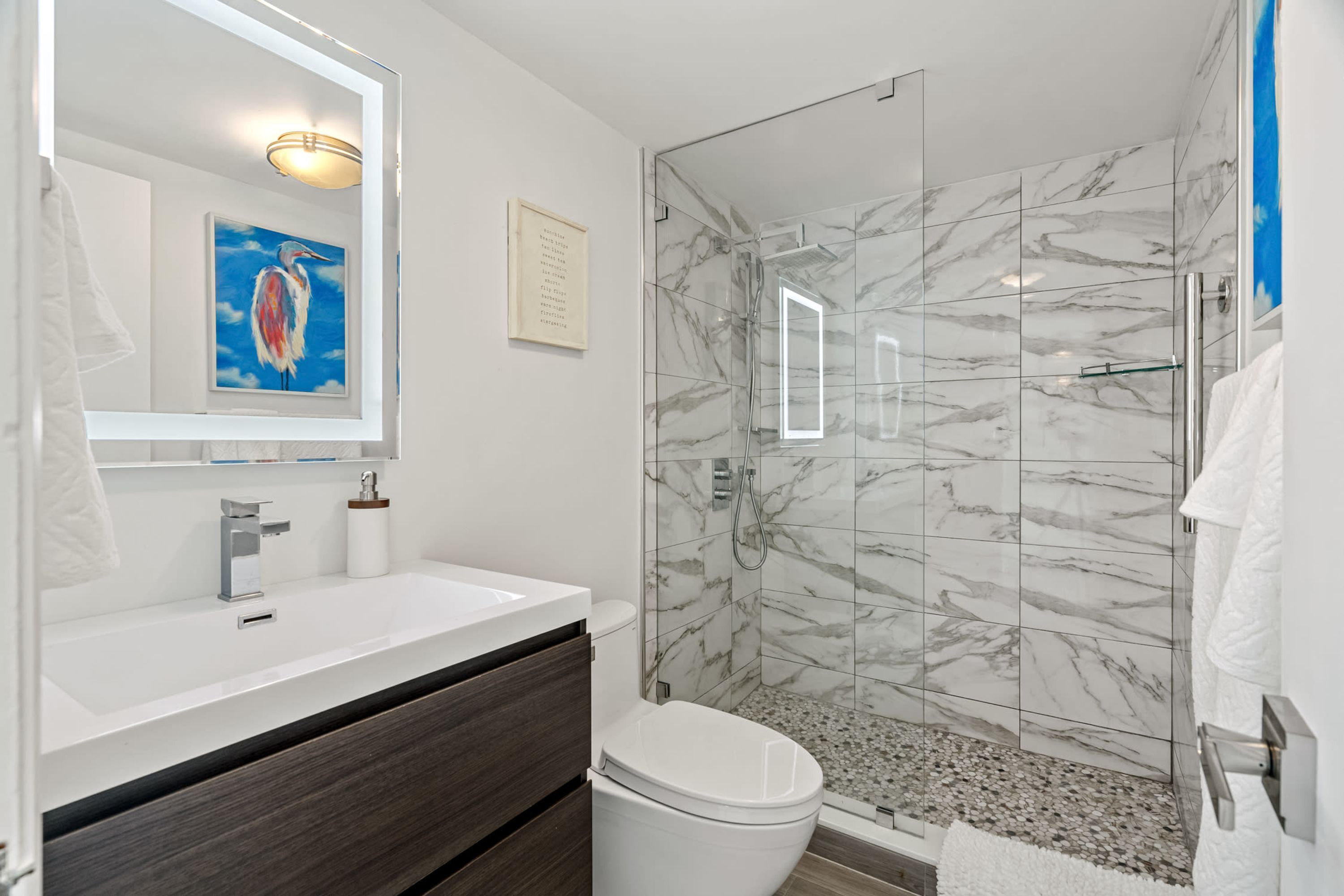 A modern bathroom with a white sink, dark wooden vanity, a white toilet, and a glass-enclosed shower with marble tiles.
