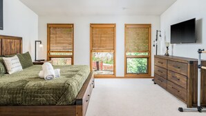 Un dormitorio con cama, cómoda, televisor y tres ventanas con persianas.