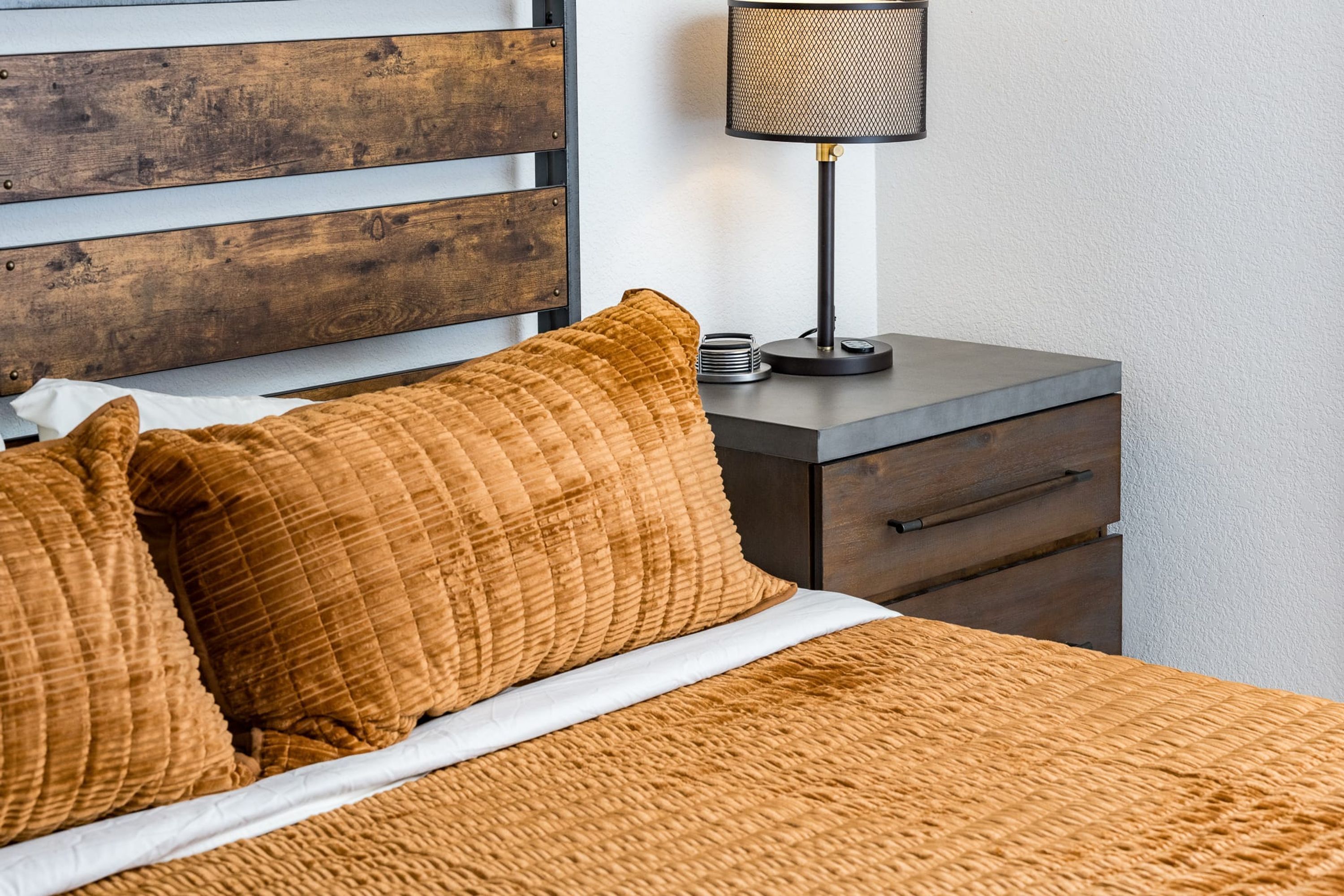 Una cama con cubrecama acolchado marrón, cabecera de madera, lámpara de noche y mesita de noche con lámpara.