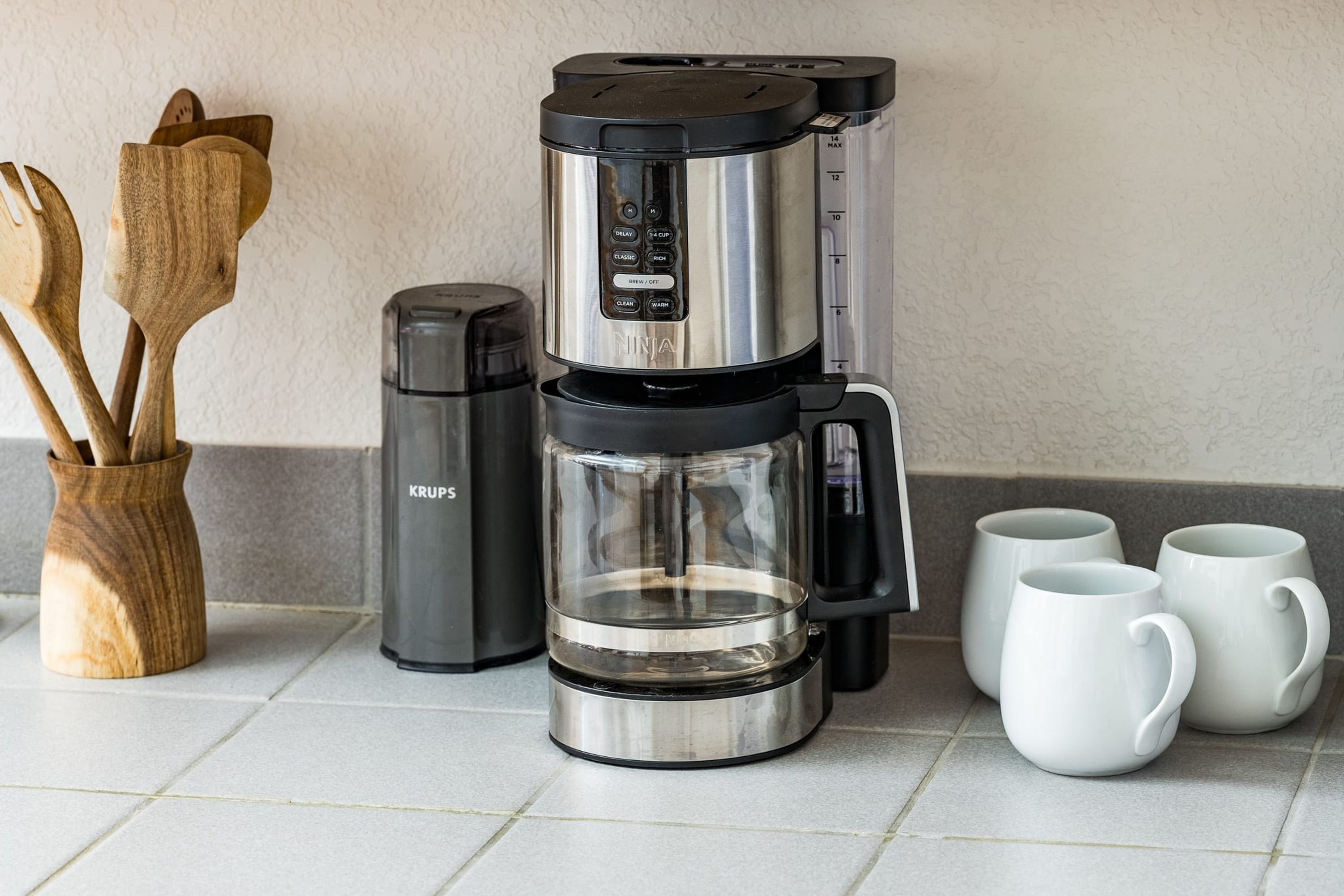Una cafetera con jarra de vidrio, un molinillo de café Krups y tres tazas blancas sobre una encimera de baldosas.
