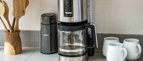 Una cafetera con jarra de vidrio, un molinillo de café Krups y tres tazas blancas sobre una encimera de baldosas.