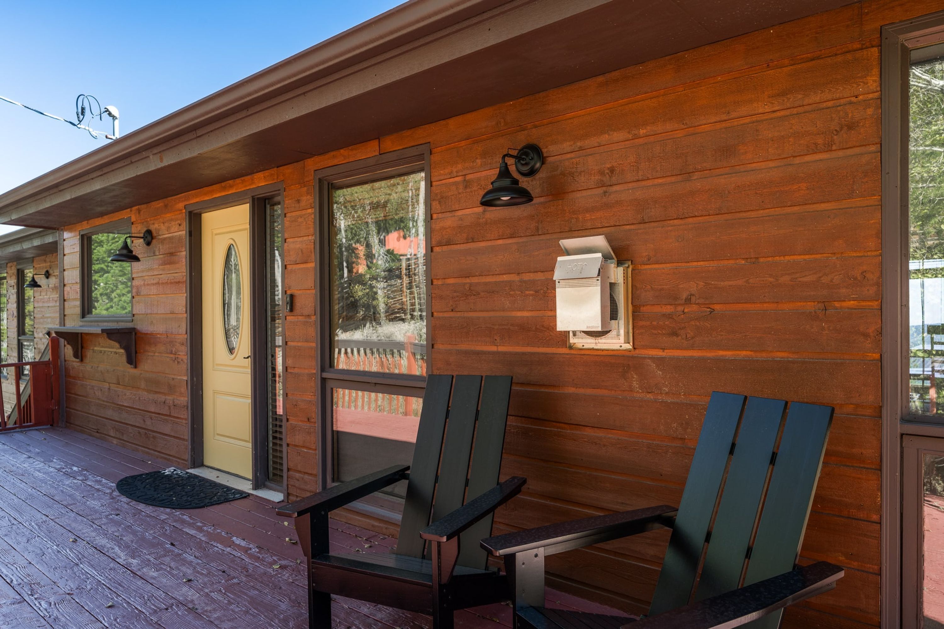 A wooden exterior with two Adirondack chairs on a porch, a door, and a wall-mounted light fixture.