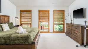 Un dormitorio con cama, cómoda, televisor y tres ventanas con persianas.