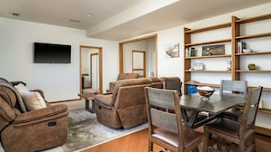 Un salon moderne comprenant un canapé, un fauteuil inclinable et une table à manger avec des chaises. Un téléviseur à écran plat est fixé au mur, et une bibliothèque contient divers objets.