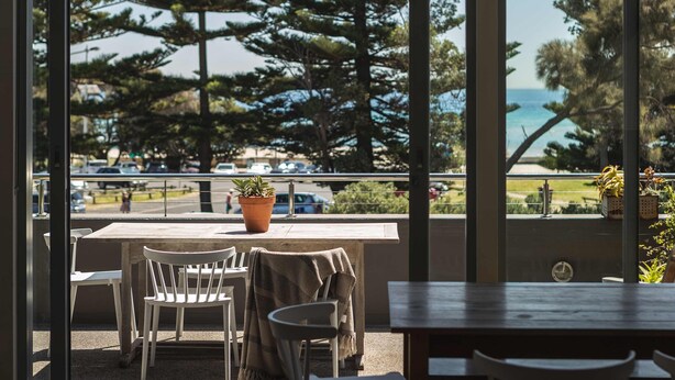 Un balcon avec une table et des chaises, donnant sur une zone côtière bordée d’arbres et où sont garées des voitures.