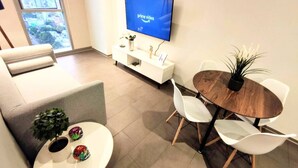 Una sala de estar moderna con un televisor de pantalla plana mostrando 'prime video', un sofá blanco, una mesa de comedor con sillas blancas y una planta en maceta.