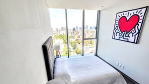 Un dormitorio pequeño con vista a la ciudad, una cama, una pared blanca y un cuadro enmarcado.