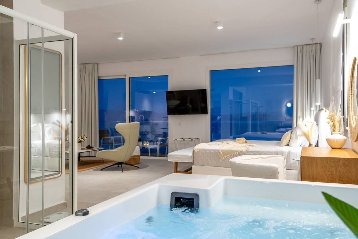 A modern hotel room with a large window, a bathtub, a bed, a chair, and a small table with a vase.