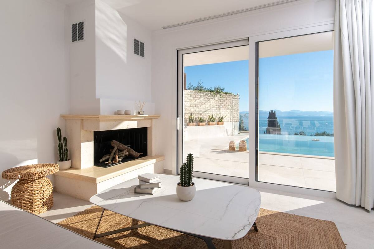 A modern living room with a fireplace, a large window with a view of a pool and mountains, and a balcony with a stone wall.