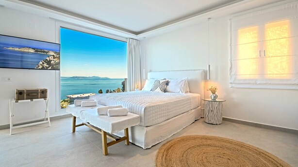 A modern bedroom with a large bed, a bench, a small table with a vase, and a view of the sea. - Achillion Luxury Corfu Villa (Corfu)