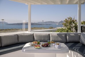 Un espace repas extérieur moderne, avec une vue panoramique sur l’océan, des sièges confortables et une variété d’aliments pour le petit-déjeuner disposés sur la table.