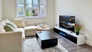 A modern living room with a sofa, a coffee table, a TV on a stand, and a window with a view of buildings. - 2 Bed Apartment - Sleeps 4, Balcony, Pets Ok (London)