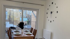 A dining area with a wooden table set for four, a large wall clock, and a view through a glass door to the outside. - 2 Bed Apartment - Sleeps 4, Balcony, Pets Ok (London)