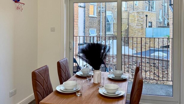 A dining table set for four with white plates and bowls, a vase with black feathers, and a view of a brick building through a glass door. - 2 Bed Apartment - Sleeps 4, Balcony, Pets Ok (London)