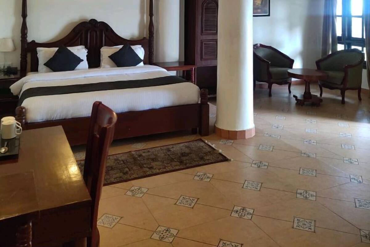A hotel room with a large bed, a wooden table, two chairs, a decorative rug, and a ceiling fan.