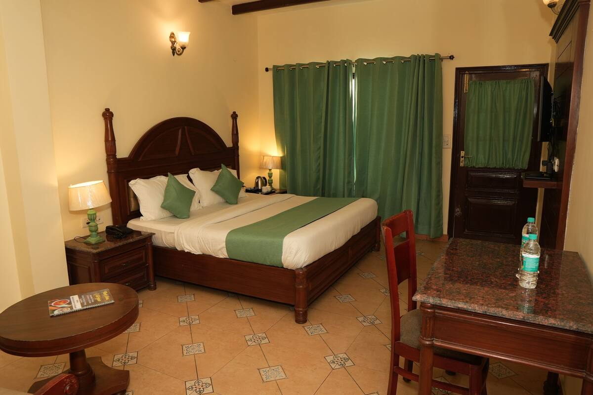 A hotel room with a wooden bed, green curtains, a nightstand with a lamp, a small table, and a chair.