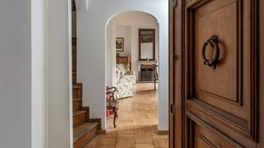 Una puerta de madera con manija de latón, una escalera que conduce a una sala de estar con chimenea y un sillón estampado con motivos florales.