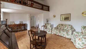 Amplia sala de estar con un piano de madera, un comedor con sillas y una mesa, un sofá floral y una escalera.