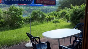 Un espace extérieur aménagé avec une table et des chaises, une enseigne indiquant « BAD DÜRHEIMER », et une vue sur un lac entouré de verdure.