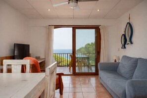 A living room with a sofa, a dining table with chairs, a television, and a balcony with a view of the sea. - Apartament amb Espectaculars Vistes al Mediterrani (Palafrugell)