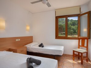 A hotel room with two beds, a wooden headboard, a desk, a chair, a window with a view of greenery, and a ceiling fan. - Apartament amb Espectaculars Vistes al Mediterrani (Palafrugell)
