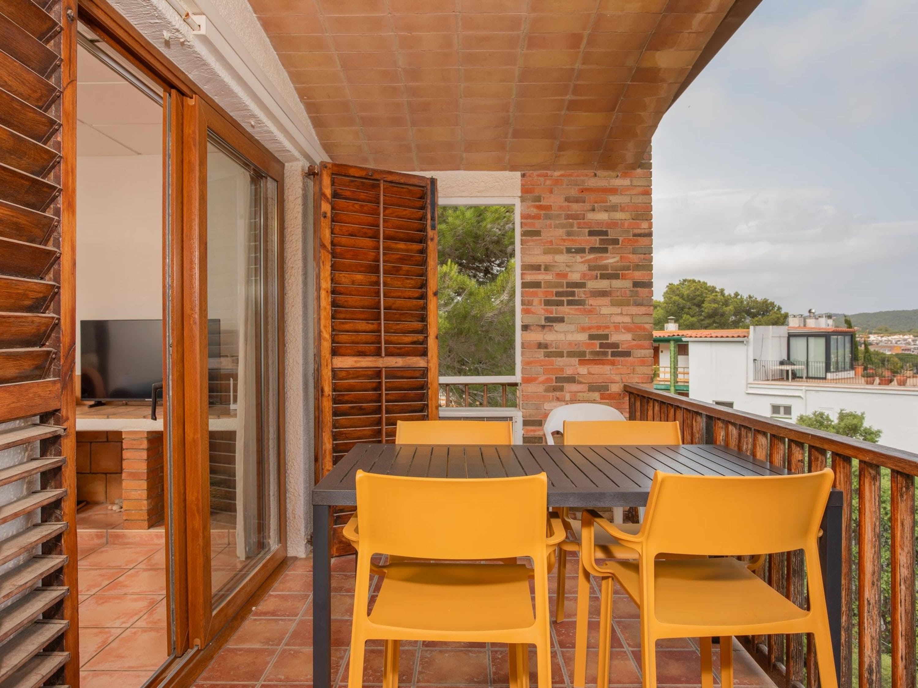 A balcony with a wooden table and yellow chairs, a TV, and a brick wall.