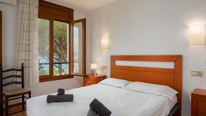 A bedroom with a bed, a chair, a nightstand, a lamp, and a window with curtains. - Apartament amb Espectaculars Vistes al Mediterrani (Palafrugell)