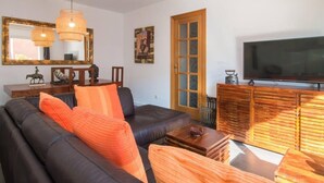 Un salon avec un canapé en cuir noir, des coussins orange, une table basse en bois, un téléviseur posé sur un meuble en bois et un grand miroir accroché au mur.