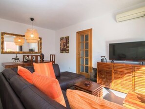 Un salon avec un canapé en cuir noir, des coussins orange, une table basse en bois, un téléviseur posé sur un meuble en bois et un grand miroir accroché au mur.