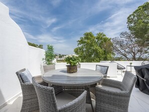 A round table with wicker chairs, a potted plant, and a view of trees and buildings in the background.