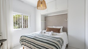 A modern bedroom with a large bed, a hanging rattan light fixture, and a view of trees through a window.