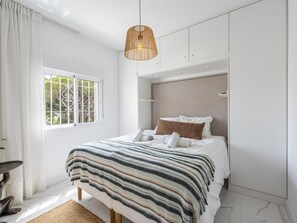 A modern bedroom with a large bed, a hanging rattan light fixture, and a view of trees through a window.