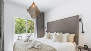 A modern bedroom with a large bed, a hanging light fixture, a view of greenery outside, and a small table with a lamp. - Contemporary new Apartment With 2 Terraces (Marbella)