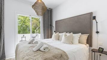 Un dormitorio moderno con una cama grande, una lámpara colgante, vistas al exterior con vegetación y una pequeña mesa con lámpara.