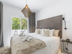 Een moderne slaapkamer met een groot bed, een hanglamp, uitzicht op groen en een klein tafeltje met een lamp.