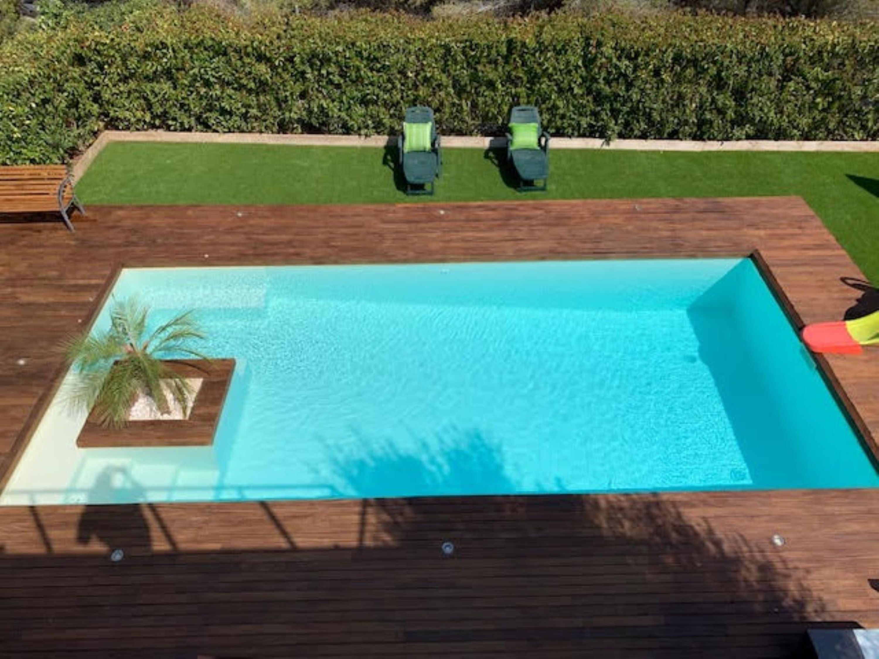 A rectangular swimming pool with a wooden deck, a palm tree, and two green lawnmowers.