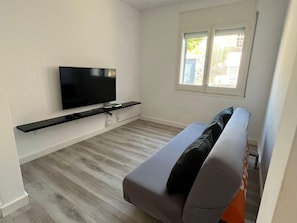 A modern living room with a flat-screen TV mounted on the wall, a grey sofa with a black cushion, and a window with blinds. - Villa Independiente con Piscina y Vistas (Sant Pere de Ribes)