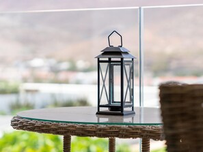 A round glass table with a woven wicker chair, a black lantern, and a view of a body of water with buildings in the background.
