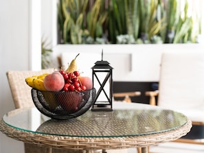 A wicker table with a glass top, a black metal fruit bowl containing bananas, cherries, and a peach, and a black lantern.