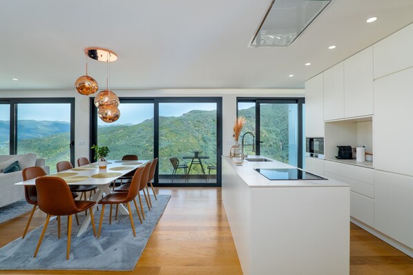 A modern kitchen with large windows, a dining area with a table and chairs, and a view of the mountains.