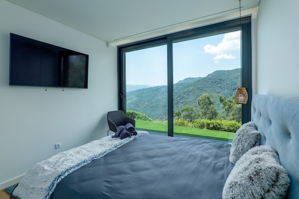 A bedroom with a large bed, a TV mounted on the wall, and a view of a mountainous landscape through a large window.