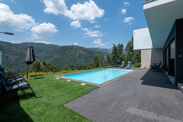 A modern outdoor pool area with a clear view of the mountains, surrounded by a wooden deck and a grassy lawn.