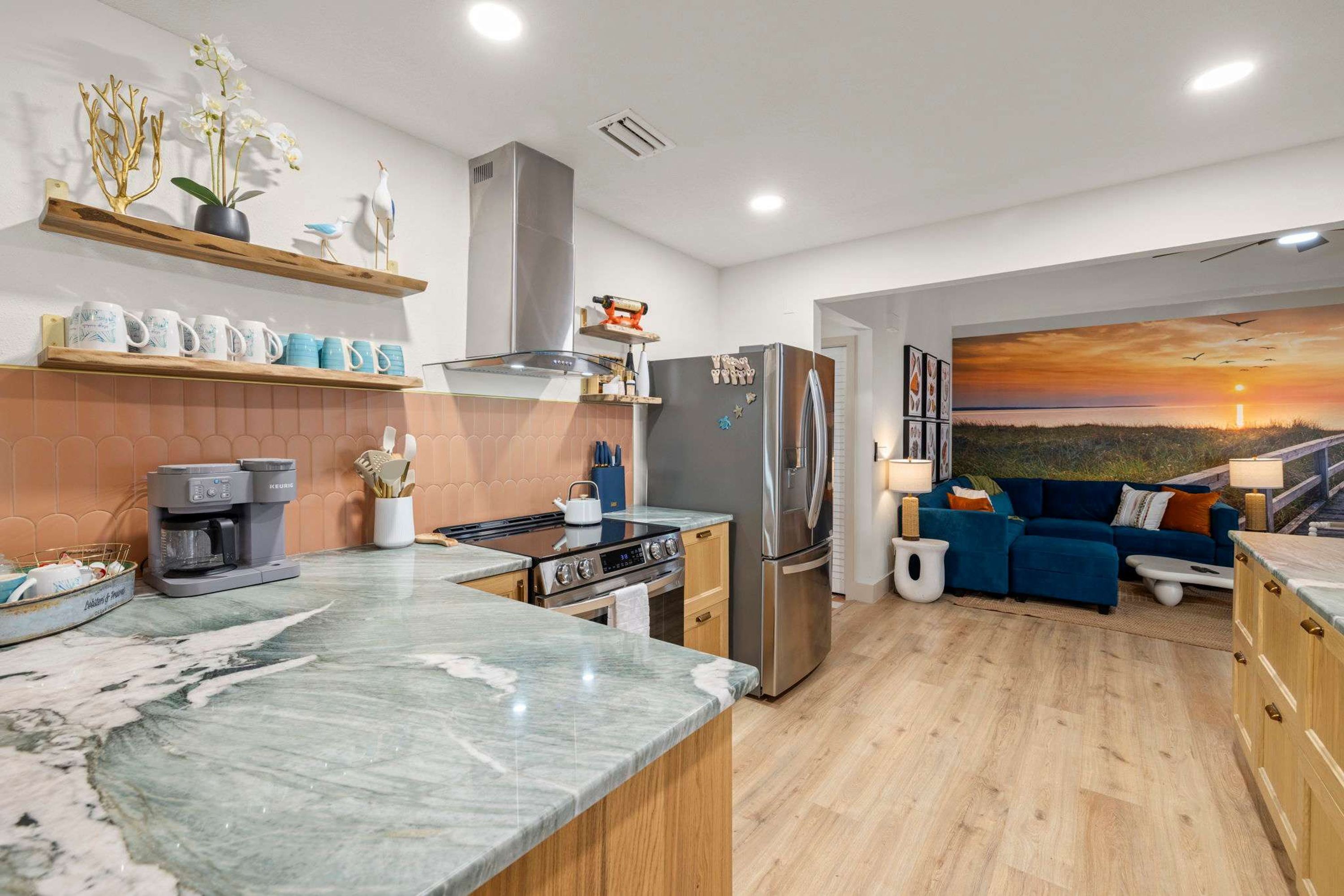 Una cocina moderna con encimera de mármol, electrodomésticos de acero inoxidable y una zona de comedor con sofá azul y un cuadro de un atardecer.