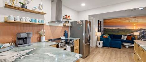 Una cocina moderna con encimera de mármol, electrodomésticos de acero inoxidable y una zona de comedor con sofá azul y un cuadro de un atardecer.