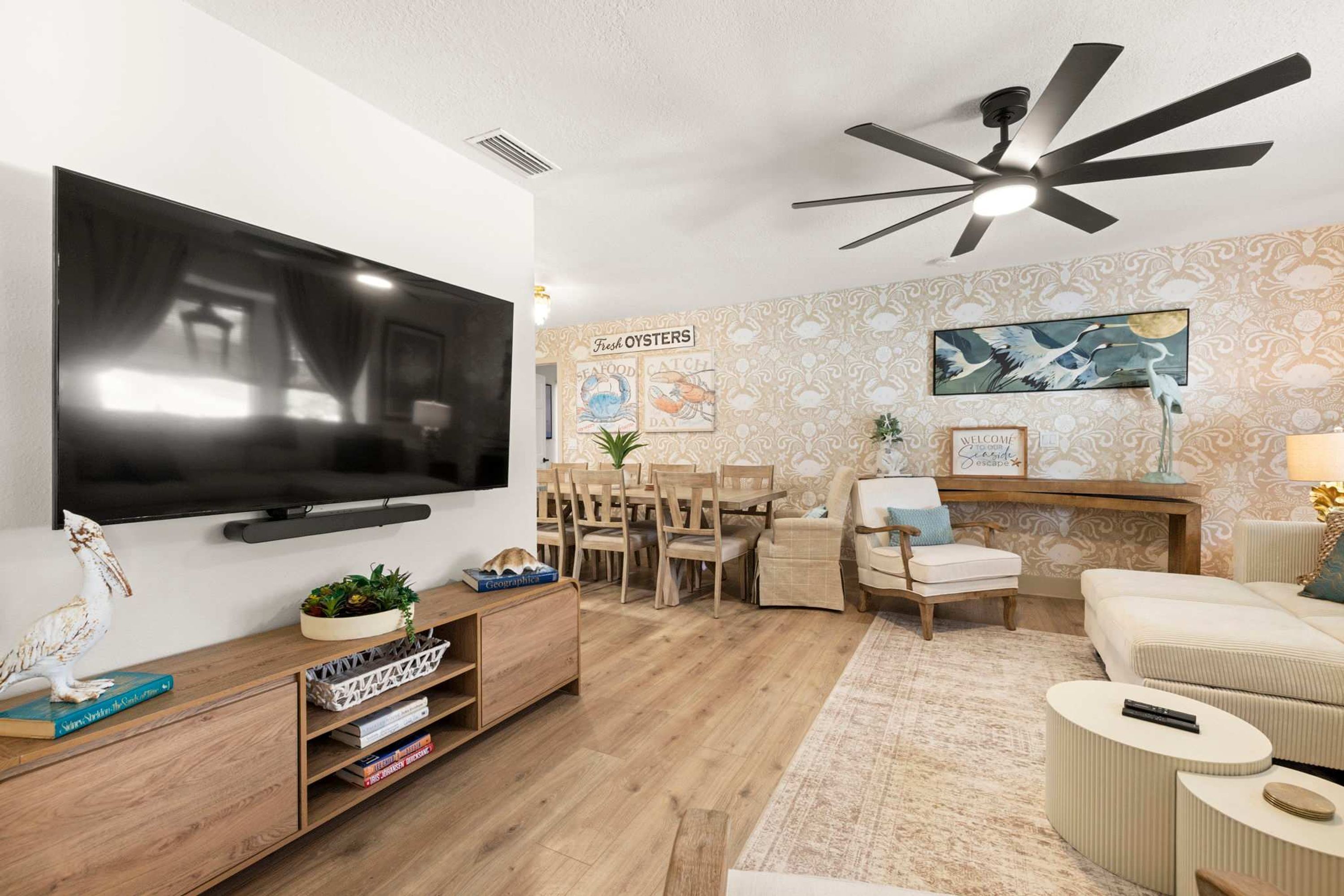 Un salon moderne doté d’un téléviseur à écran plat, de meubles en bois, d’un ventilateur de plafond et d’un mur orné d’œuvres d’art sur le thème des fruits de mer.