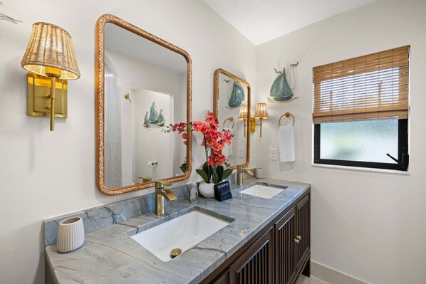 A bathroom with two sinks, a large mirror, wall sconces, and a window with blinds.