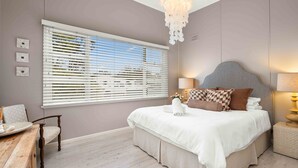 Un dormitorio con cama, una silla, una mesita de noche, una lámpara y una ventana con persianas.